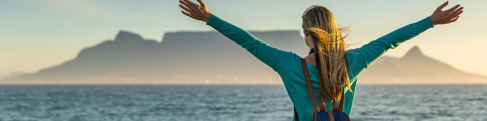 A woman stands with her arms stretched on the beach with a view of Table Mountain in Cape Town, which can be visited with a cheap holiday package from Flight Centre.